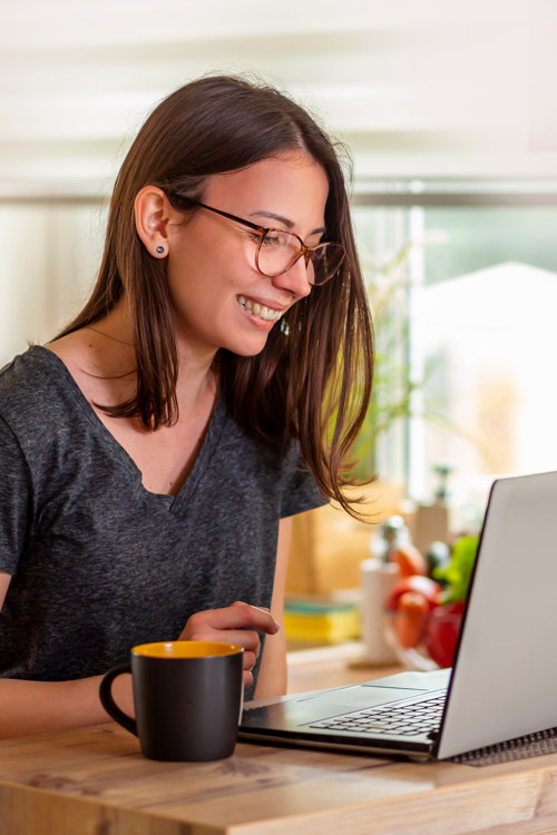 A woman at a kitchen table looking at a laptop with a warm cup of coffee